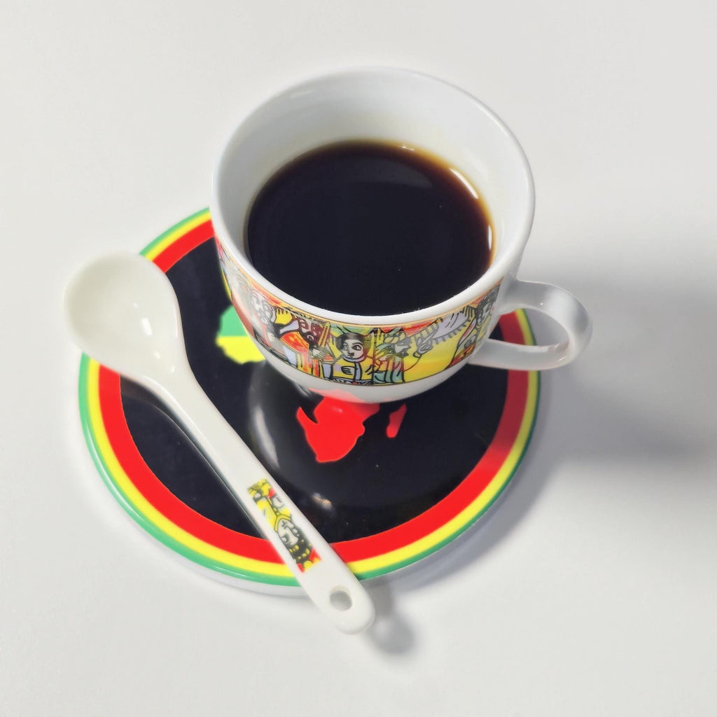 A white ceramic cup filled with black coffee sits on a matching saucer and spoon. The cup and saucer feature a colorful illustrated border, and the saucer has bold black, red, yellow, and green concentric rings on a white surface.