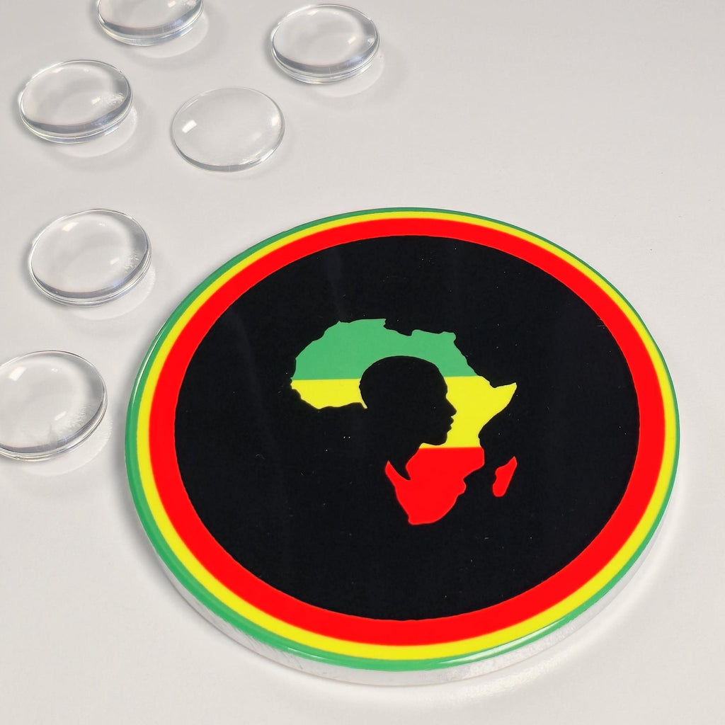 A round clear acrylic or glass coaster with a black center featuring a silhouette of Africa filled with the Rastafarian flag colors (green, yellow, red) on a black background. The coaster is surrounded by a thick border of red, yellow, and green concentric rings.