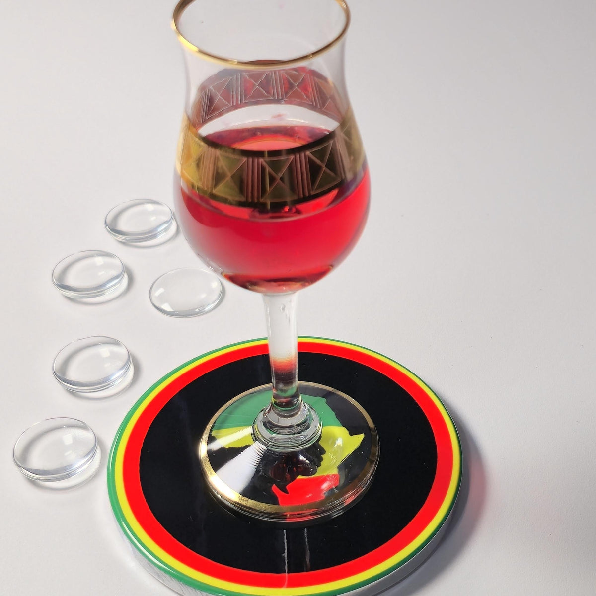 A stemmed glass with a gold-trimmed rim and geometric pattern, partially filled with red liquid, resting on a round black coaster with red, yellow, and green stripes. Five small clear oval pieces are arranged to the left on a light gray background.