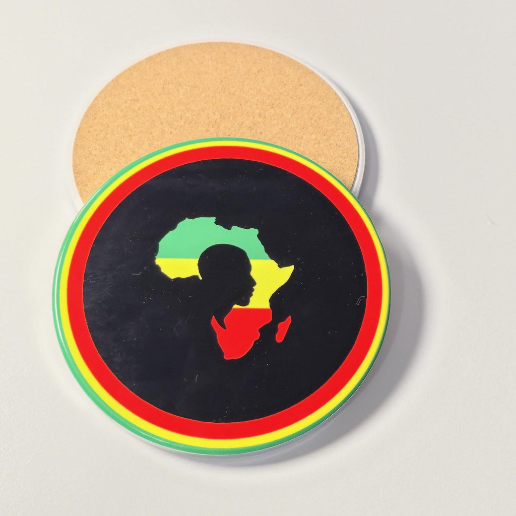 A round black coaster featuring a silhouette of Africa filled with the green, yellow, and red flag colors, surrounded by concentric red, yellow, and green rings. A second coaster showing the natural cork backing is partially visible behind it on a light gray surface.