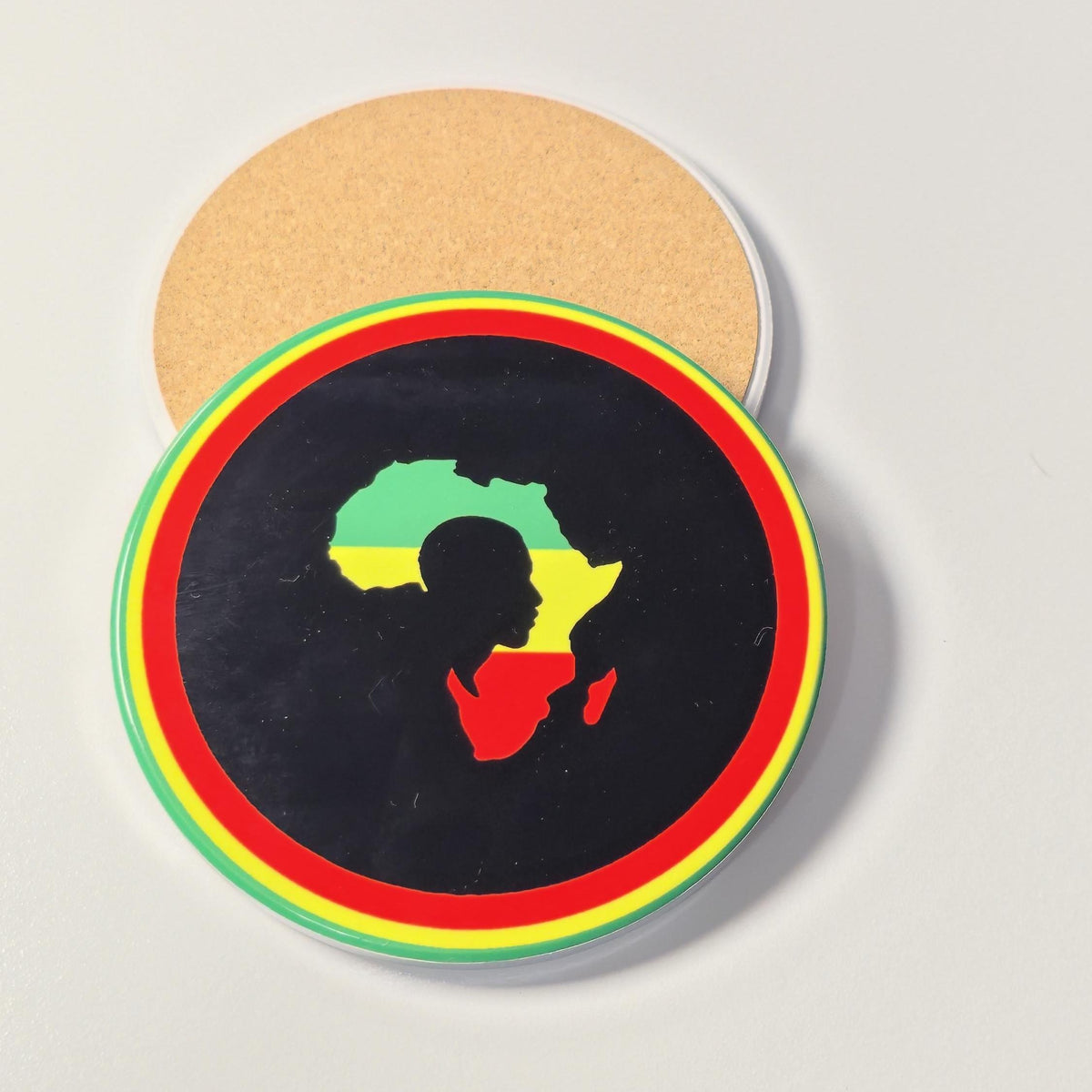 A round black coaster featuring a silhouette of Africa filled with the green, yellow, and red flag colors, surrounded by concentric red, yellow, and green rings. A second coaster showing the natural cork backing is partially visible behind it on a light gray surface.
