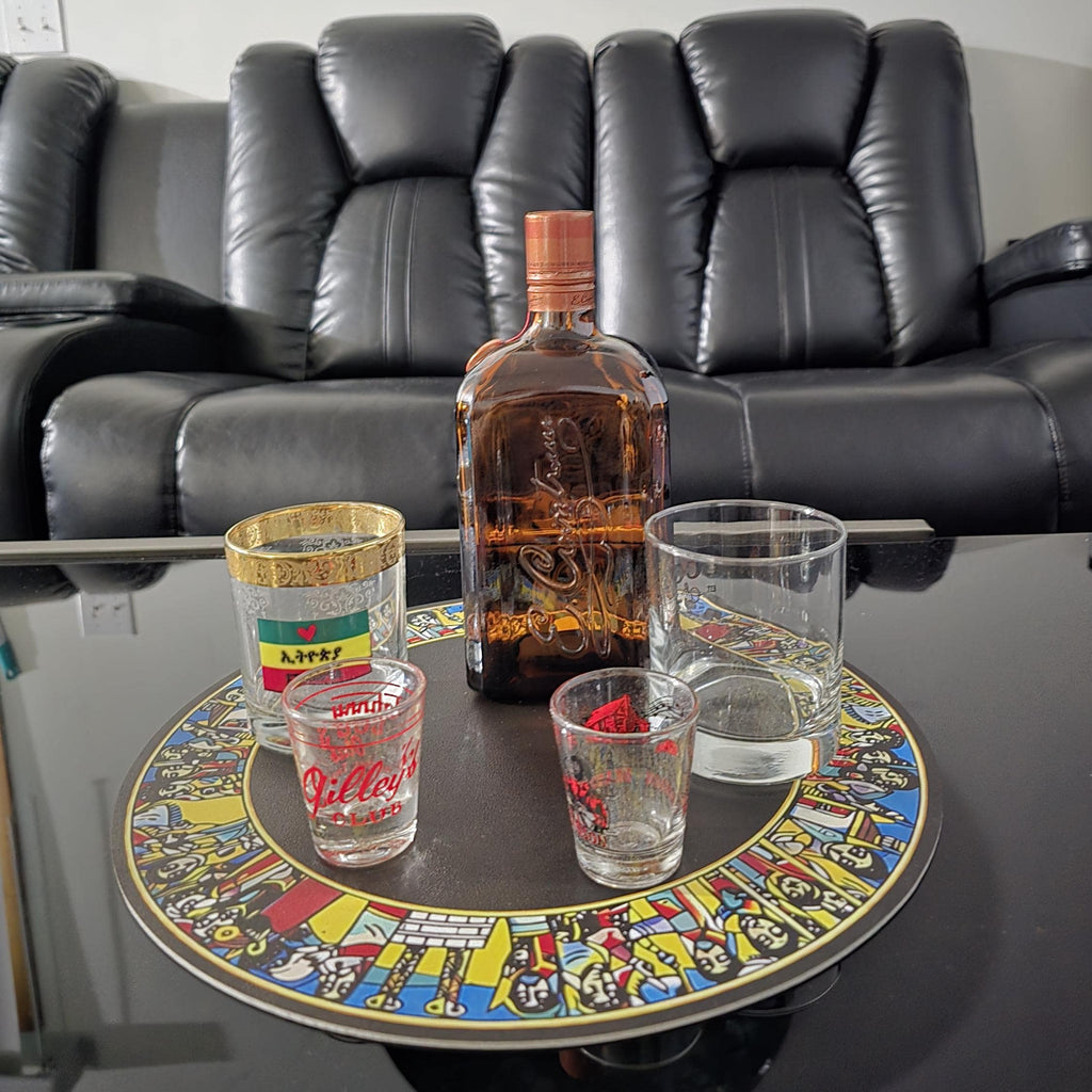A bottle of liquor with an orange cap sits on a round decorative tray with four drinking glasses, placed in front of a black leather recliner sofa in a living room.