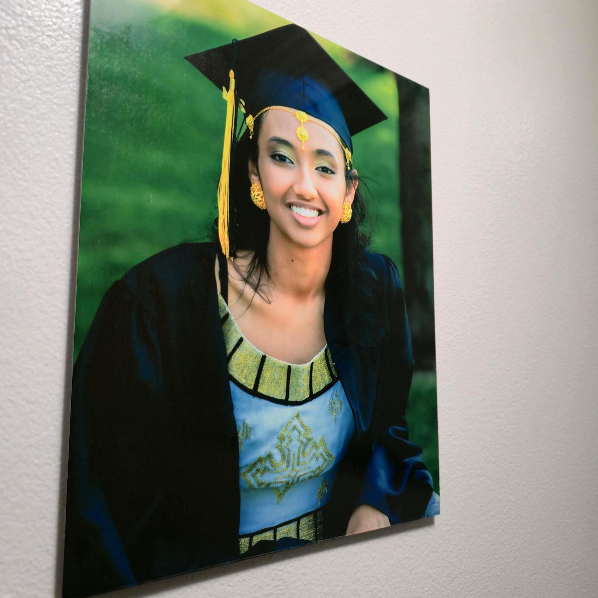 A vertical custom portrait photo print of a woman in a graduation cap and gown, displayed on a textured off-white wall.