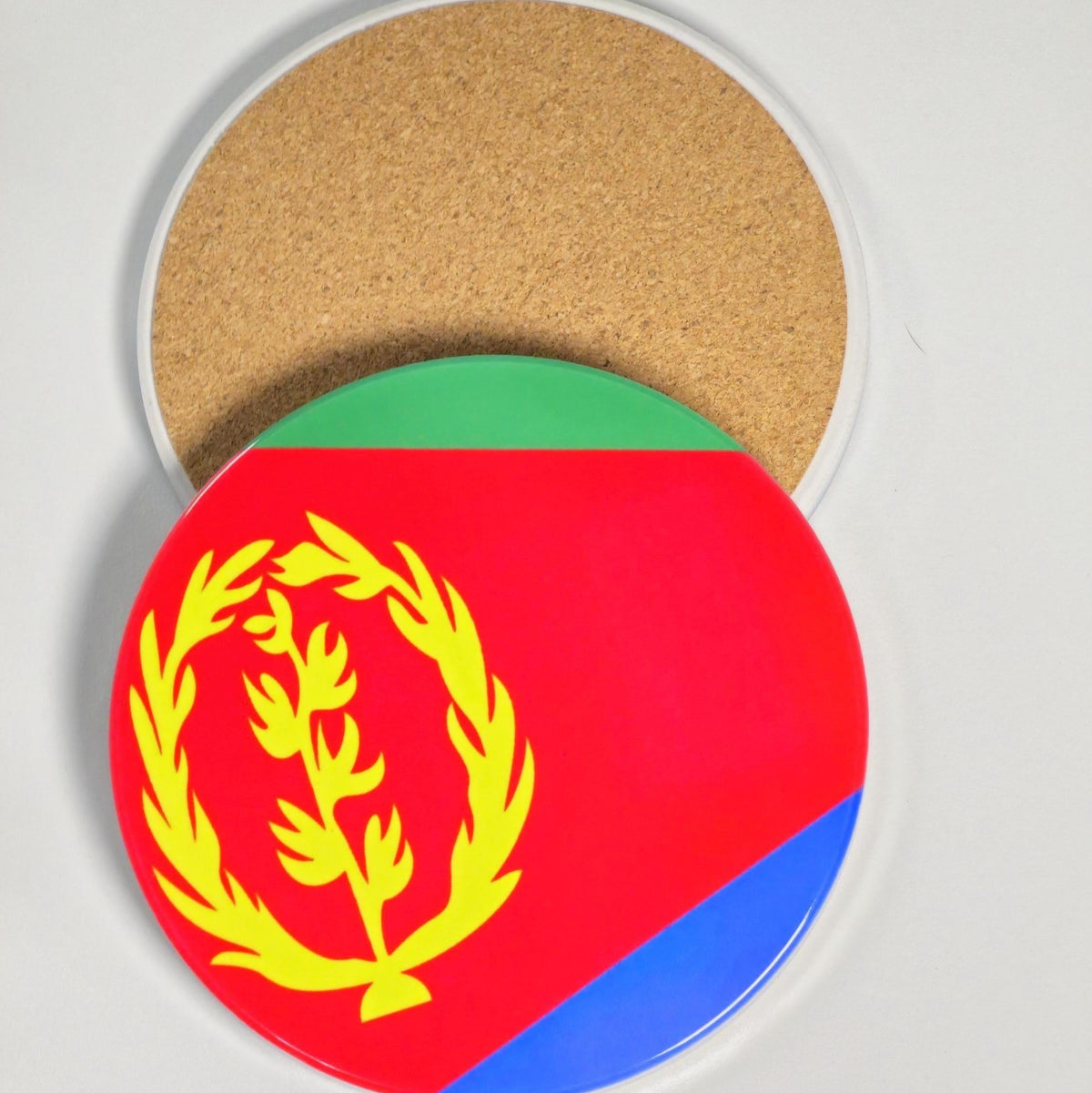 A round coaster with a printed design of the Eritrea flag, shown partially slid off a larger cork coaster on a light gray background.