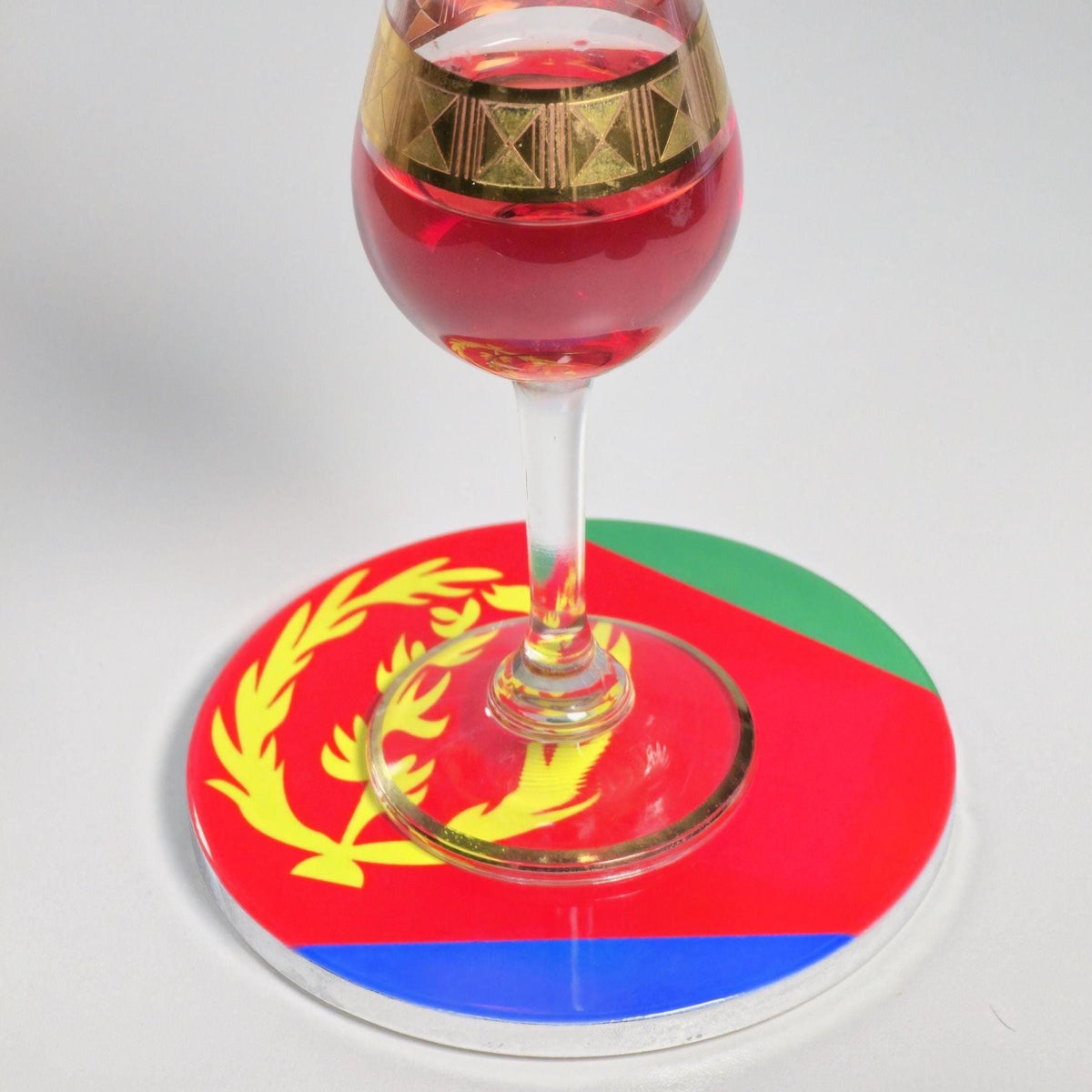 A clear stemmed glass containing red liquid, decorated with a gold band near the bowl, resting on a round clear coaster printed with the flag of Eritrea.