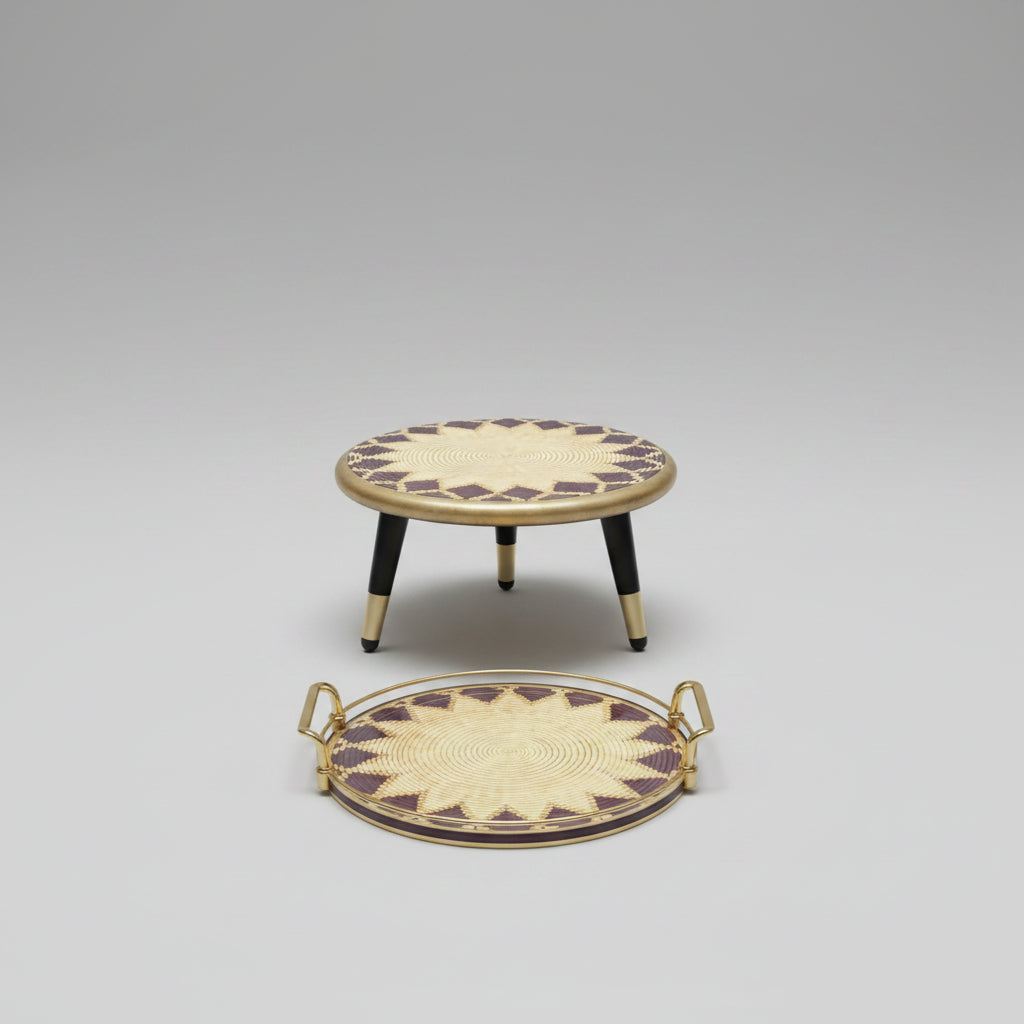 A woven round low stool and matching oval tray with gold-toned handles, featuring a cream and dark brown geometric starburst pattern, displayed on a wood floor against a light gray wall.