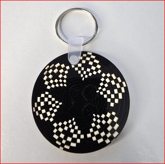 A circular black keychain featuring a white and black checkerboard spiral pattern, attached to a silver metal split ring with a clear plastic connector, shown on a light gray background.