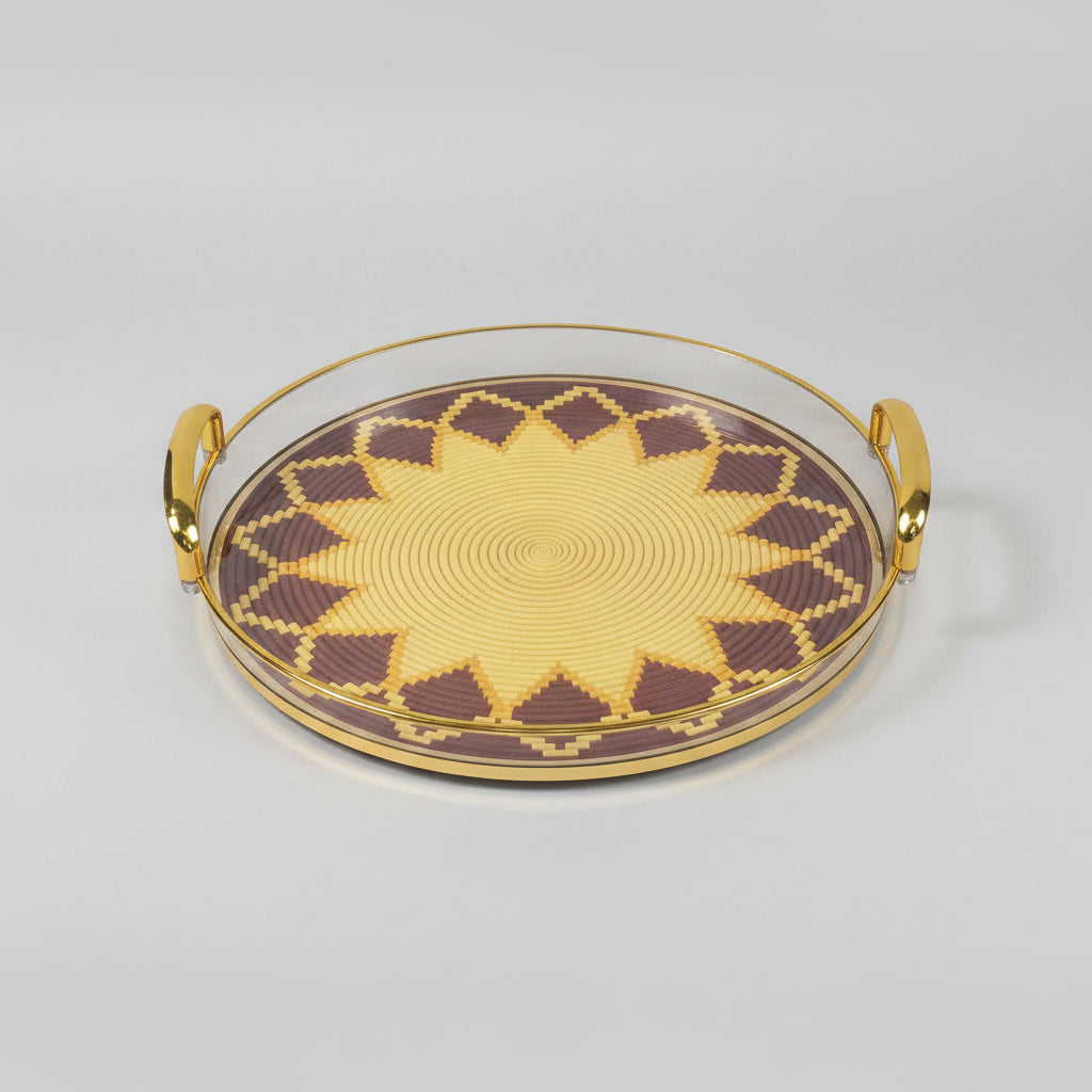 A round decorative tray with a geometric starburst pattern in yellow and dark brown, featuring a gold-toned rim and side handles, set on a black marble surface.