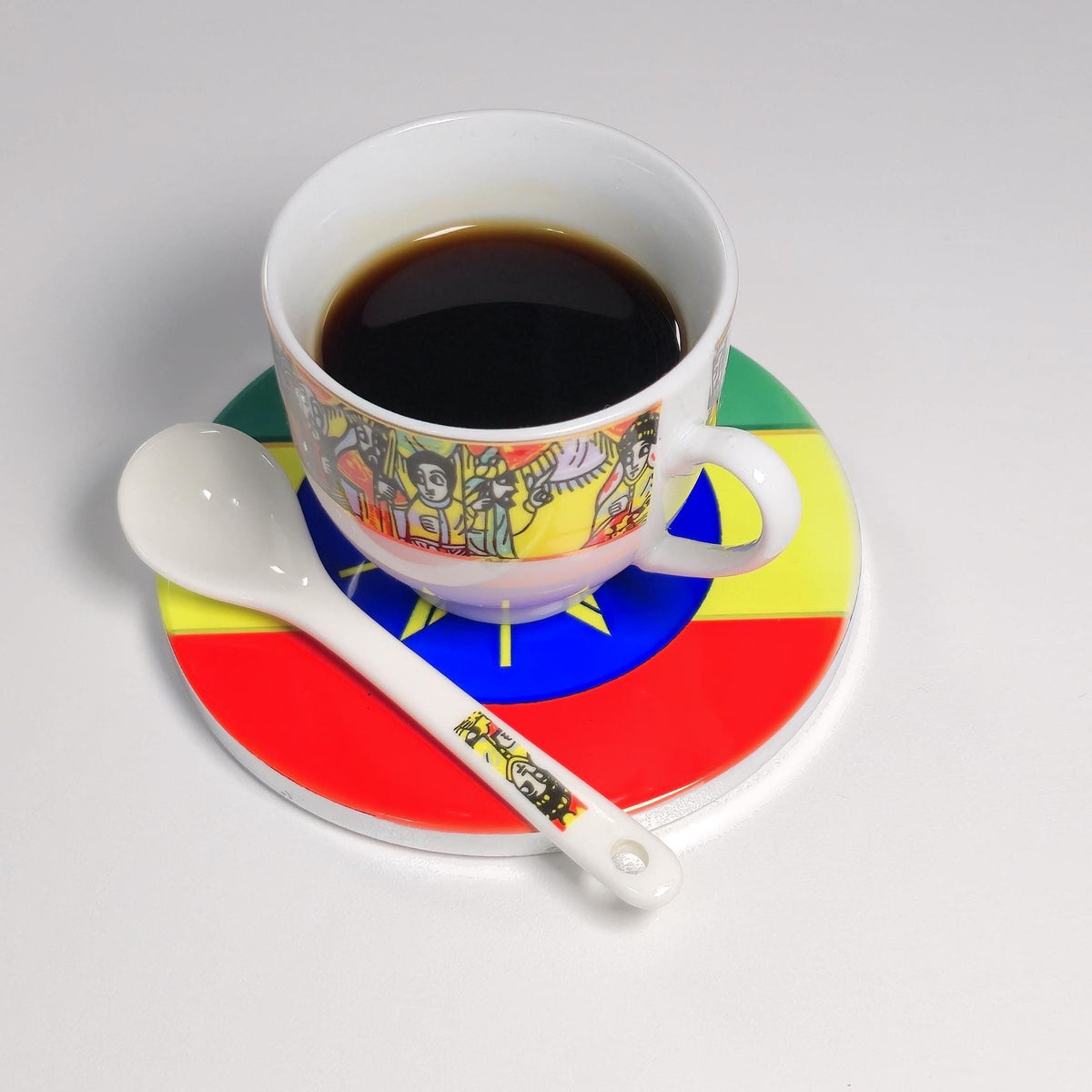 A white ceramic coffee cup filled with black coffee sits on a matching saucer and spoon. The cup and saucer are decorated with a colorful illustration and a central circular motif in green, yellow, blue, and red, resembling the Ethiopian flag.