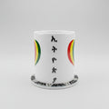 A white ceramic mug featuring the word 'HAPPY' spelled out in black Rastafarian-style letters, flanked by green, yellow, and red semicircles. The mug sits on a matching white coaster with a black and white beaded pattern, set against a neutral beige background.