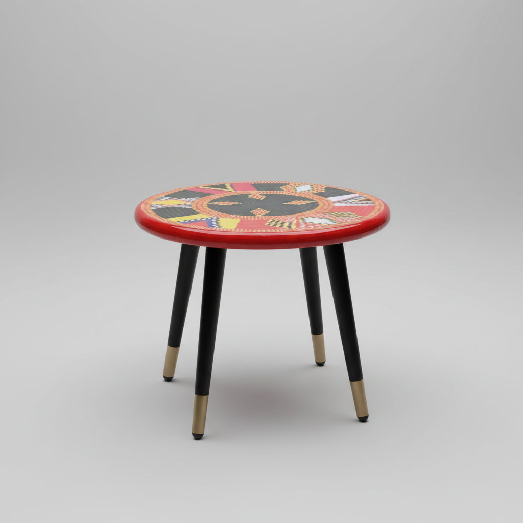 A round side table with a vibrant red top featuring a detailed geometric tribal-style pattern, supported by three black legs with gold metal caps, set on a black marble floor.