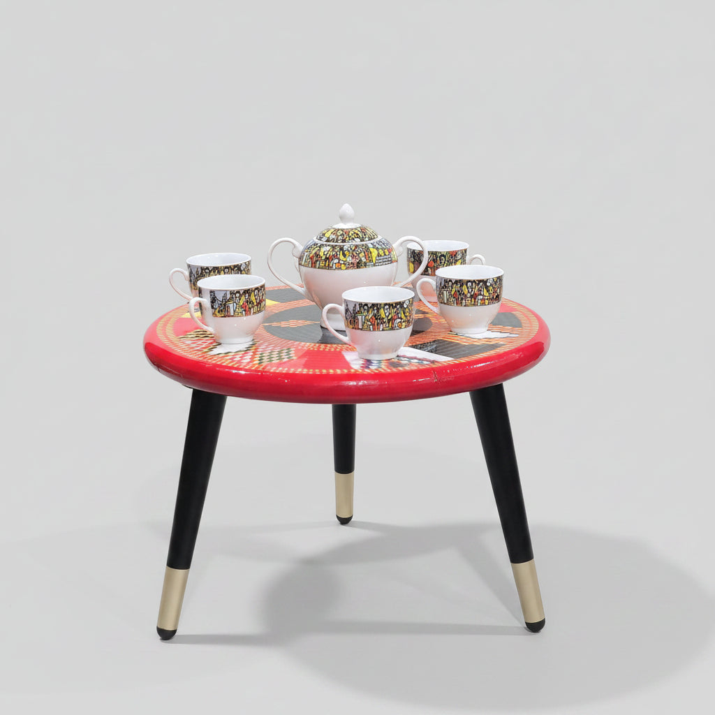 A small round table set with a red top, black legs with gold metal caps, and a colorful geometric pattern. The set includes a teapot, teapot lid, four teacups, four saucers, and a small white pedestal bowl, all arranged on a black marble-patterned floor against a light gray wall.