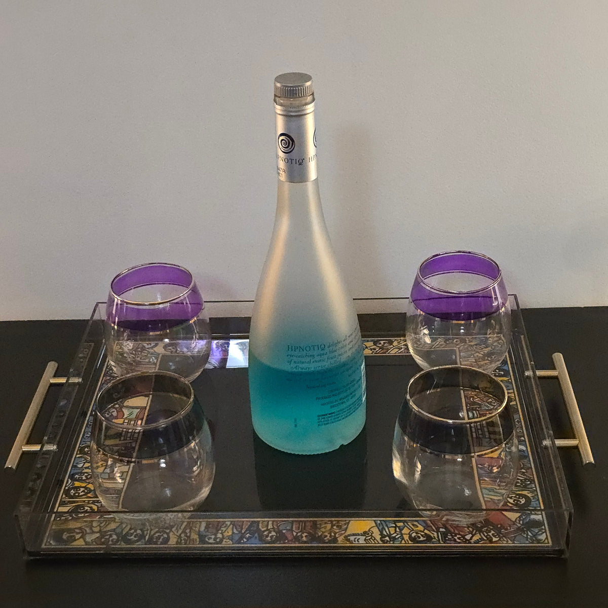 A frosted glass bottle with a blue gradient at the bottom, displayed on a decorative mirrored tray with four purple-rimmed glasses on a dark table against a plain wall.
