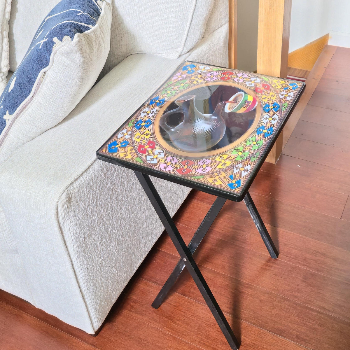 A small rectangular black metal folding side table with a colorful hand-painted mosaic top, shown next to a light beige sofa on a wooden floor.
