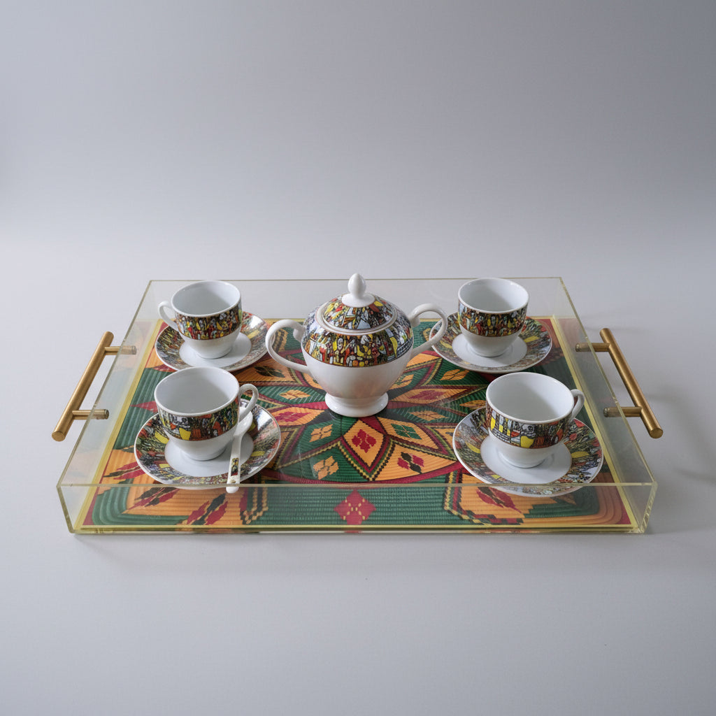 A clear acrylic serving tray with gold handles holds a colorful ceramic teapot and four matching teacups and saucers on a patterned mat, set on a black marble surface.