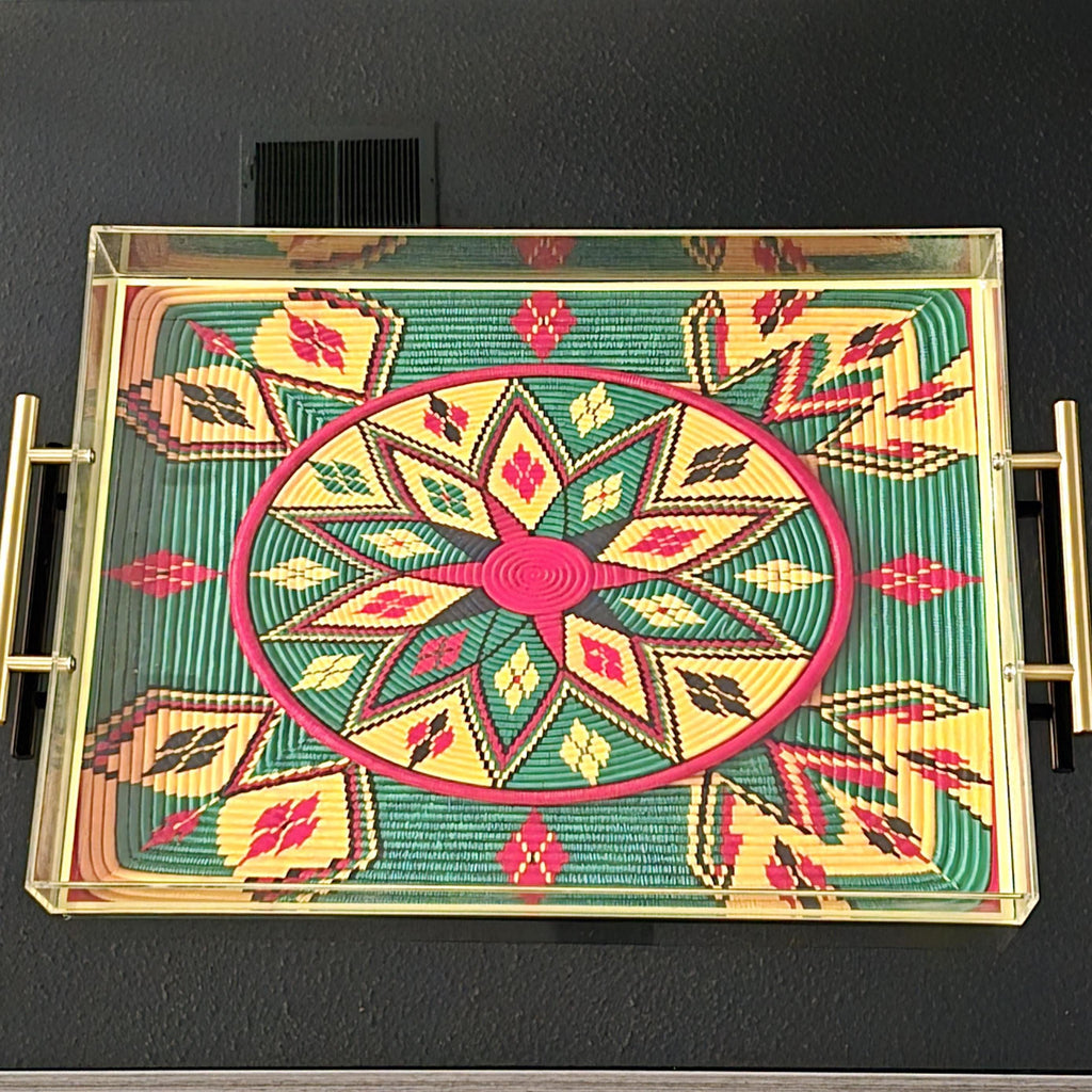 A rectangular glass-top serving tray with gold-toned metal handles, featuring a colorful Ethiopian-style geometric pattern in green, yellow, red, and black on a dark wall.