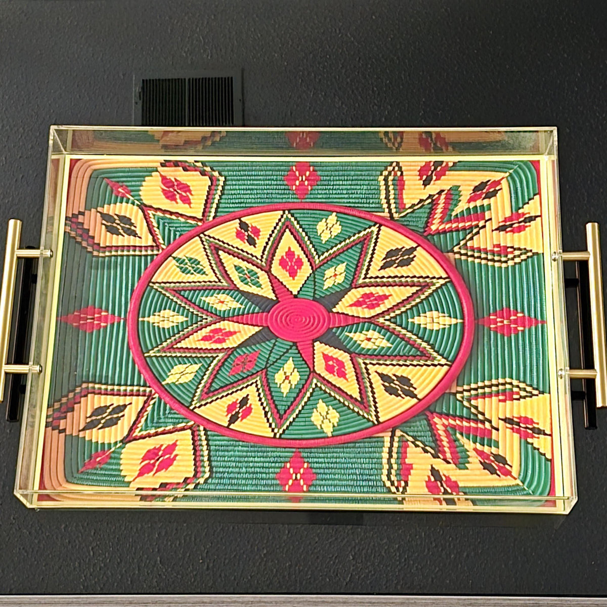 A rectangular glass-top serving tray with gold-toned metal handles, featuring a colorful Ethiopian-style geometric pattern in green, yellow, red, and black on a dark wall.