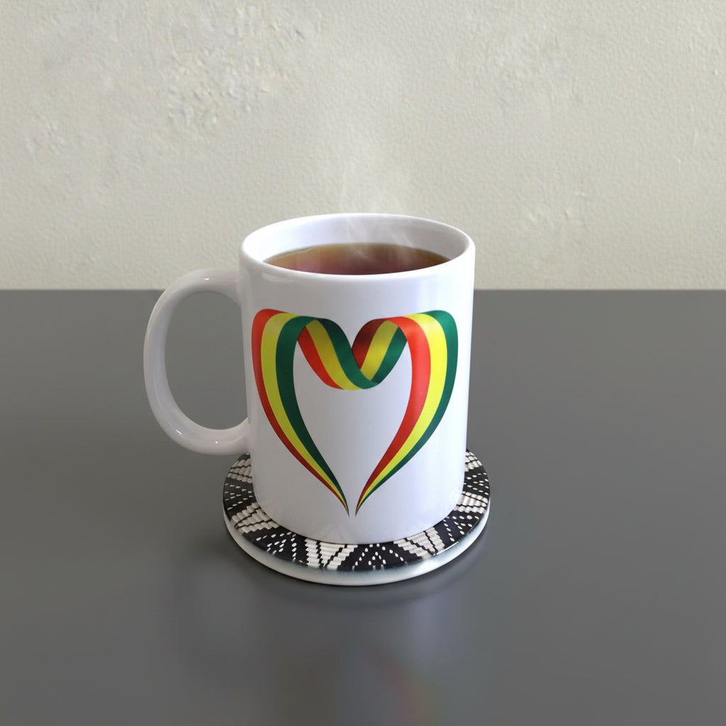 A white ceramic mug with a colorful heart design in green, yellow, and red stripes, sitting on a black and white patterned coaster on a textured beige surface.