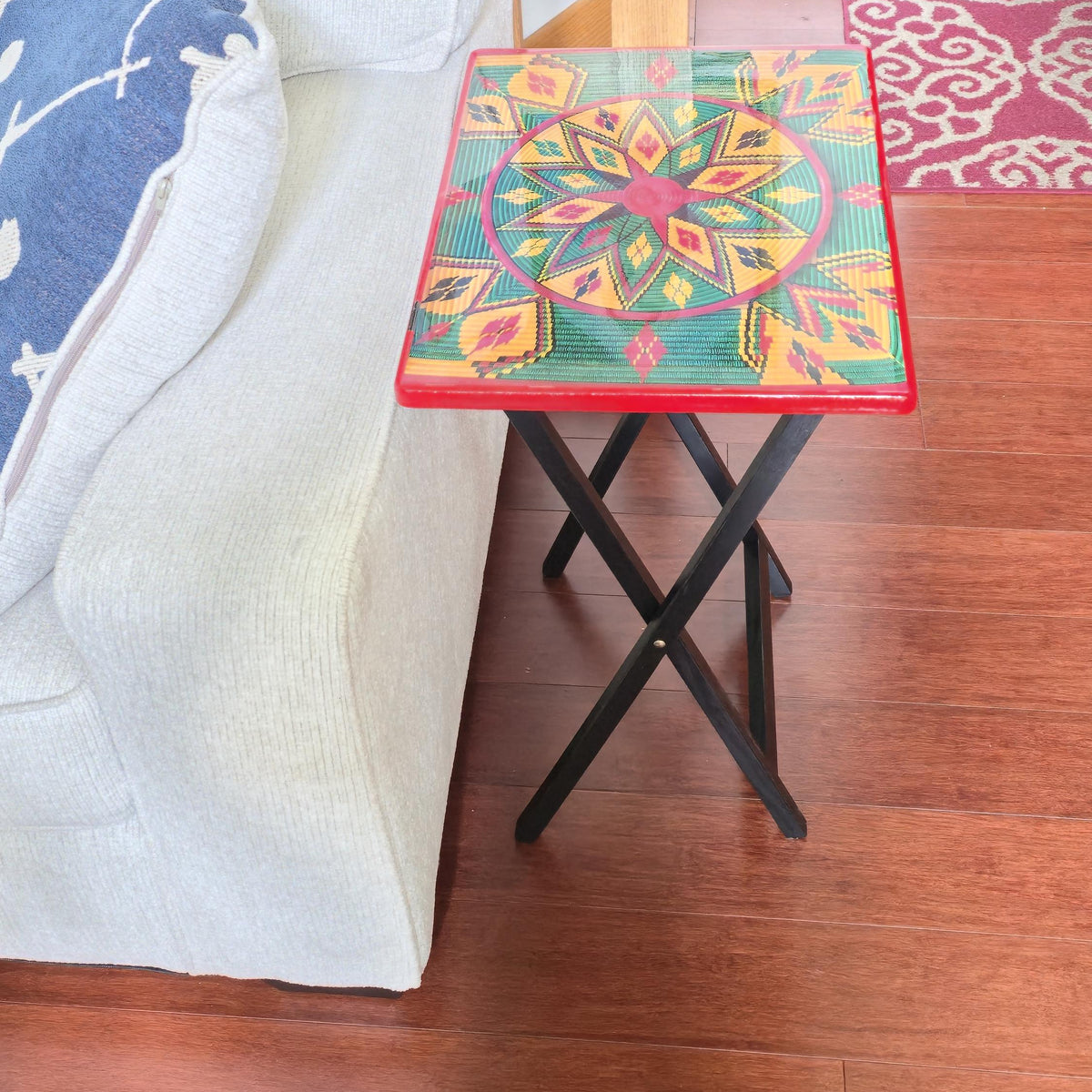 A square folding side table with a colorful geometric patterned top and black metal X-frame legs, shown next to a light-colored sofa on a wooden floor.
