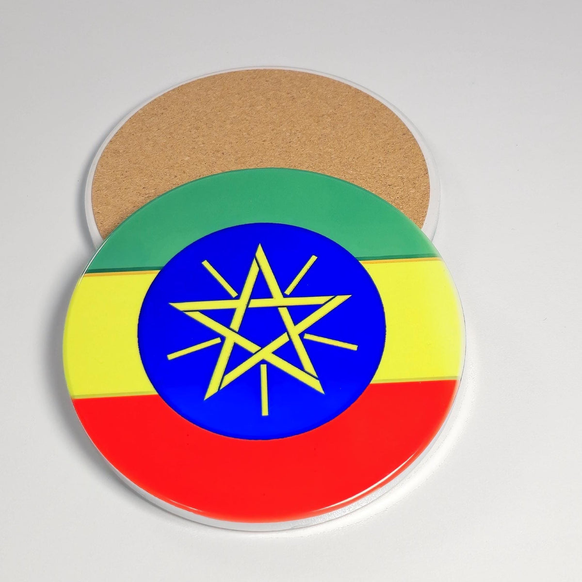 A round coaster featuring the flag of Ethiopia, with a green, yellow, and red horizontal tricolor and a blue center containing a yellow six-pointed star. A second coaster is partially visible behind it on a light gray background.