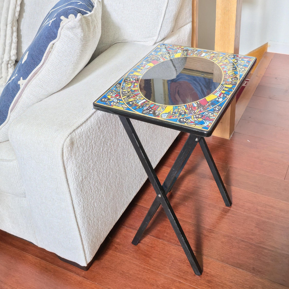 A small rectangular side table with a colorful mosaic tile top and a round mirror in the center, supported by black metal legs, positioned next to a light beige sofa on a wooden floor.