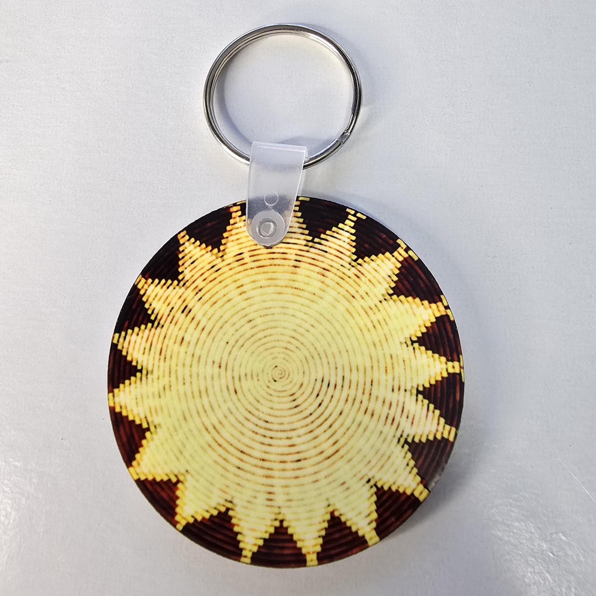 A round woven keychain featuring a sunburst pattern in natural beige and dark brown, attached to a silver metal key ring with a clear plastic connector, shown on a light gray background.