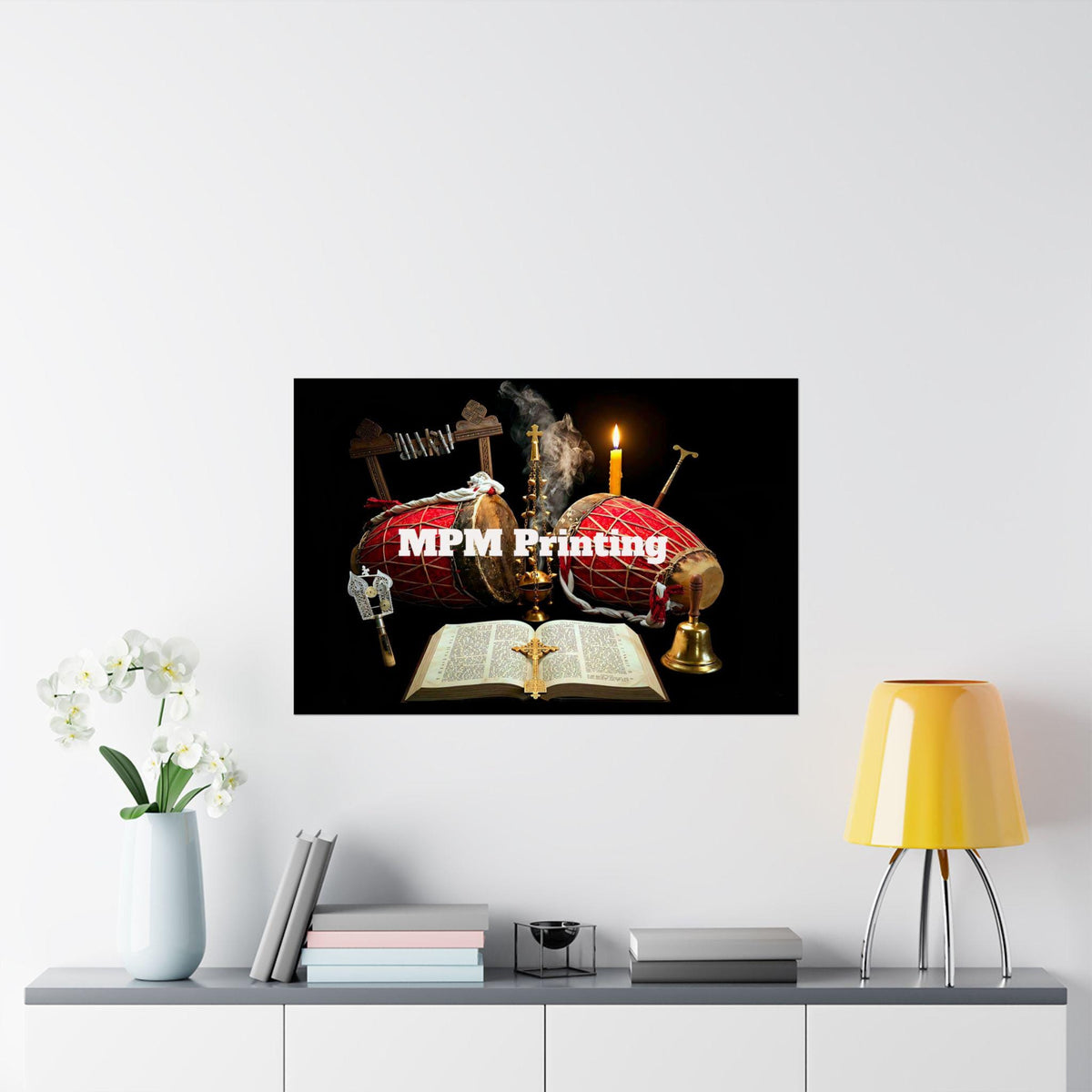 A framed-style wall print showing a black background with a red shroud, candles, an open Bible, and crosses, hung on a white wall above a shelf with flowers, books, and a yellow lamp. The print has the text 'MPM Printing' across the center.