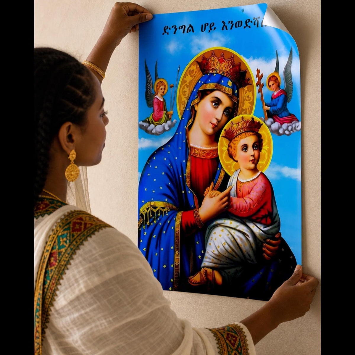 A woman in a white and gold traditional outfit hangs a colorful religious icon depicting the Virgin Mary holding the infant Jesus against a light beige wall. The icon features a bright blue sky background with Hebrew text at the top.