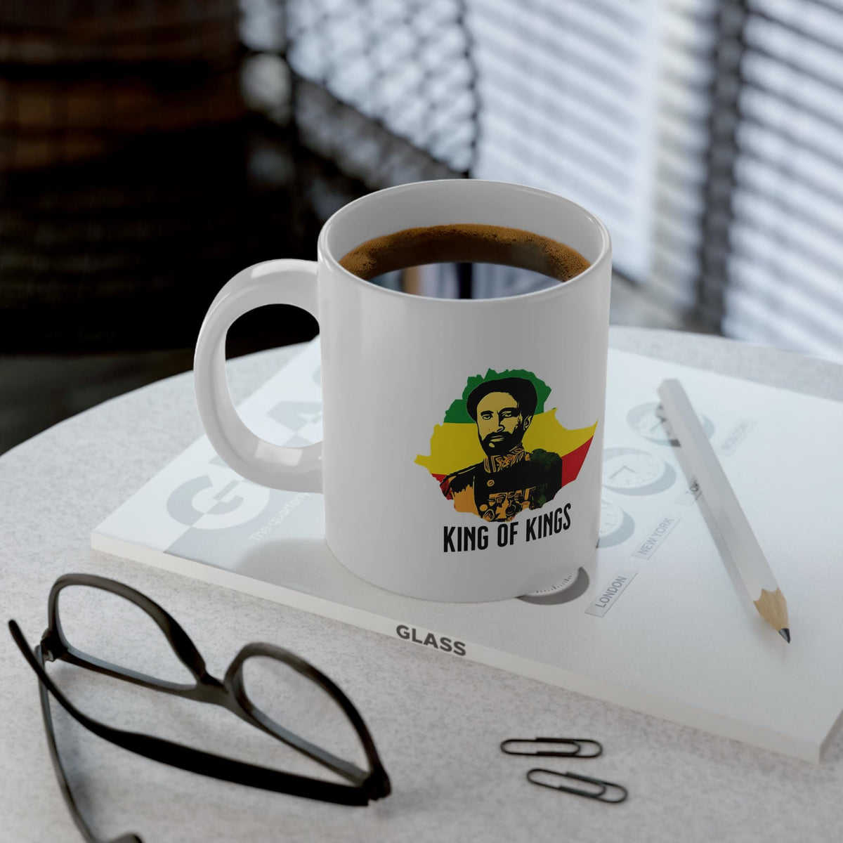 A white ceramic coffee mug filled with coffee sits on a tabletop, printed with a graphic of a man's portrait over a yellow, green, and red flag design and the text 'KING OF KINGS.' The mug is styled with eyeglasses, paper clips, a pencil, and a book in a real indoor setting with window blinds in the background.