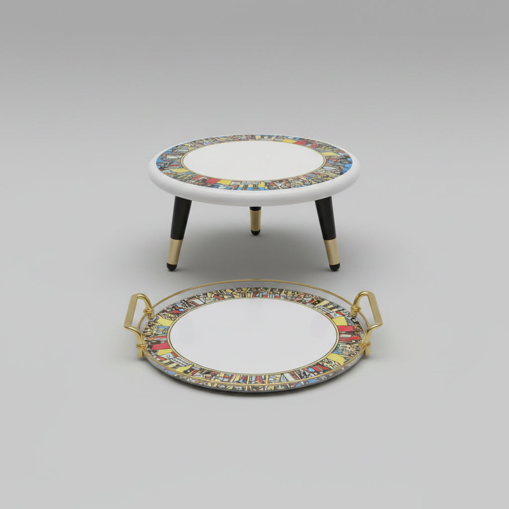 A round decorative tray with gold handles and a matching small round stool, both featuring a white center and a colorful illustrated border, placed on a wood floor against a light wall.