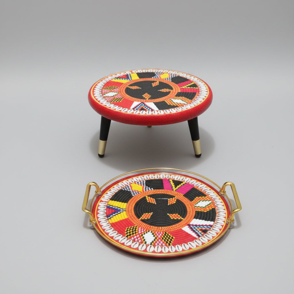 A round decorative tray with gold handles and a matching small round stool, both featuring a colorful South Asian geometric pattern in red, black, orange, yellow, pink, and white, set on a wooden floor against a white wall.