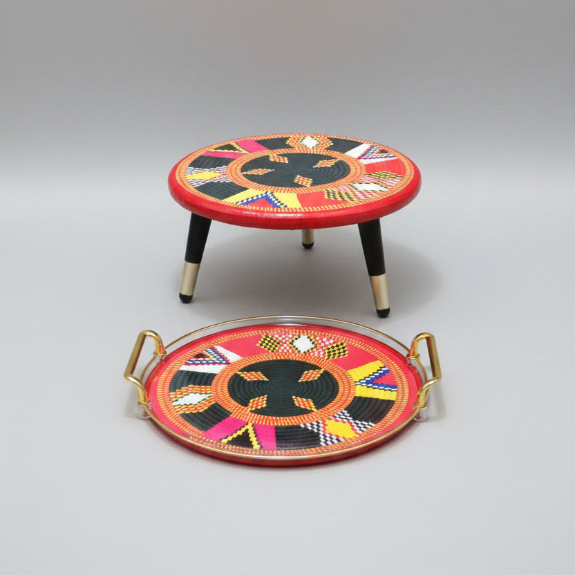 A matching set of a small round stool and a large oval tray with colorful geometric woven patterns, displayed on a wood floor against a light gray wall.