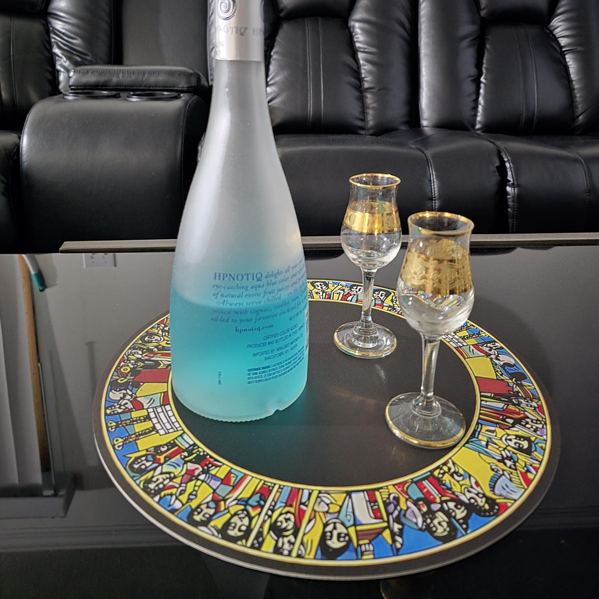 A frosted glass bottle of Bruges sparkling wine with a vibrant blue gradient at the bottom, displayed on a colorful round table next to two gold-rimmed glasses in front of a black leather recliner sofa.