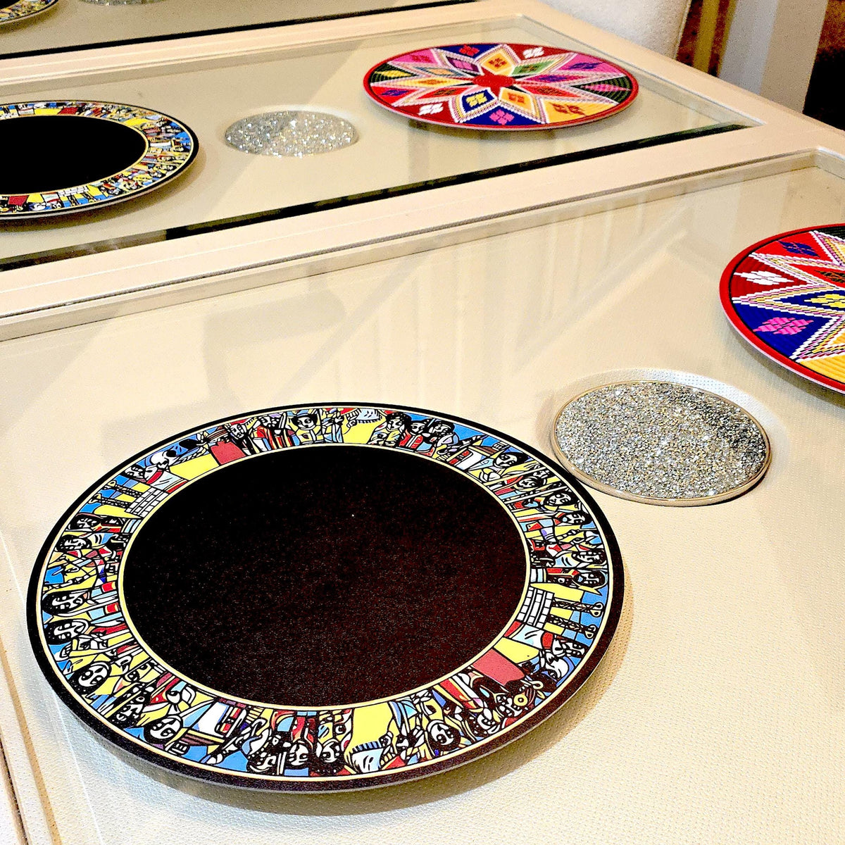 A round decorative plate with a colorful illustrated border and a dark center, displayed on a glossy cream surface with other matching decorative items nearby.