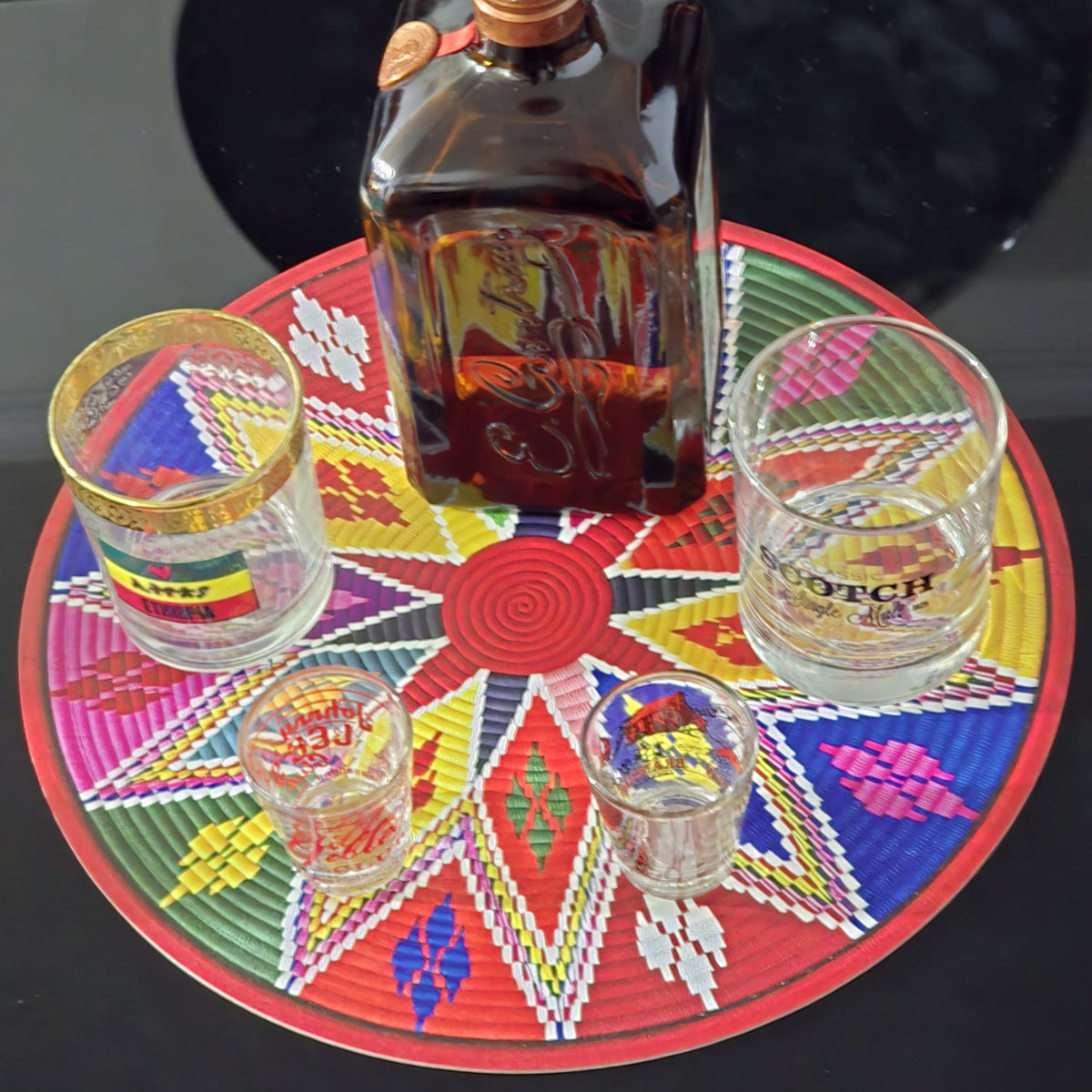 A bottle of Scotch whisky with four shot glasses arranged on a colorful circular mat, set on a glossy black surface.
