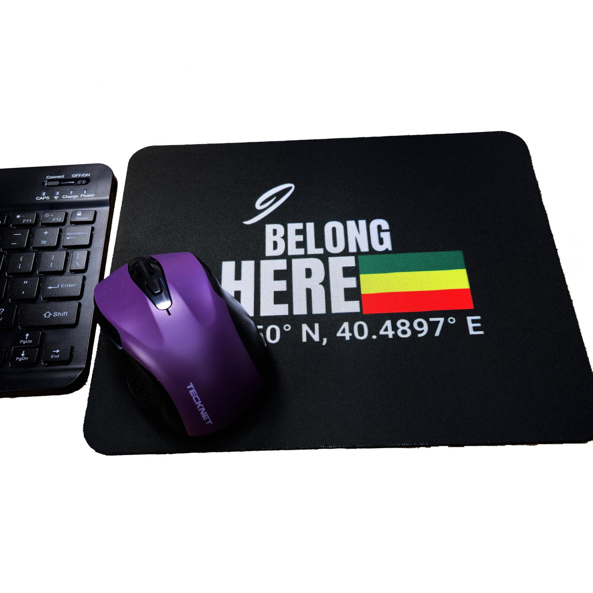 A black mouse pad with a white 'BELONG HERE' graphic, latitude/longitude coordinates, and a small green-yellow-red flag, shown on a white background next to a keyboard and a purple computer mouse.