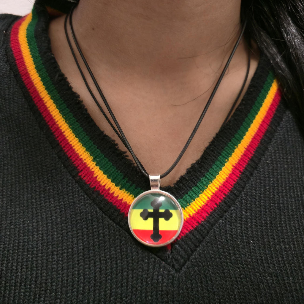 A close-up of a person wearing a black necklace with a round pendant featuring a black cross over the Rastafarian flag colors of green, yellow, and red, styled on a black V-neck sweater.