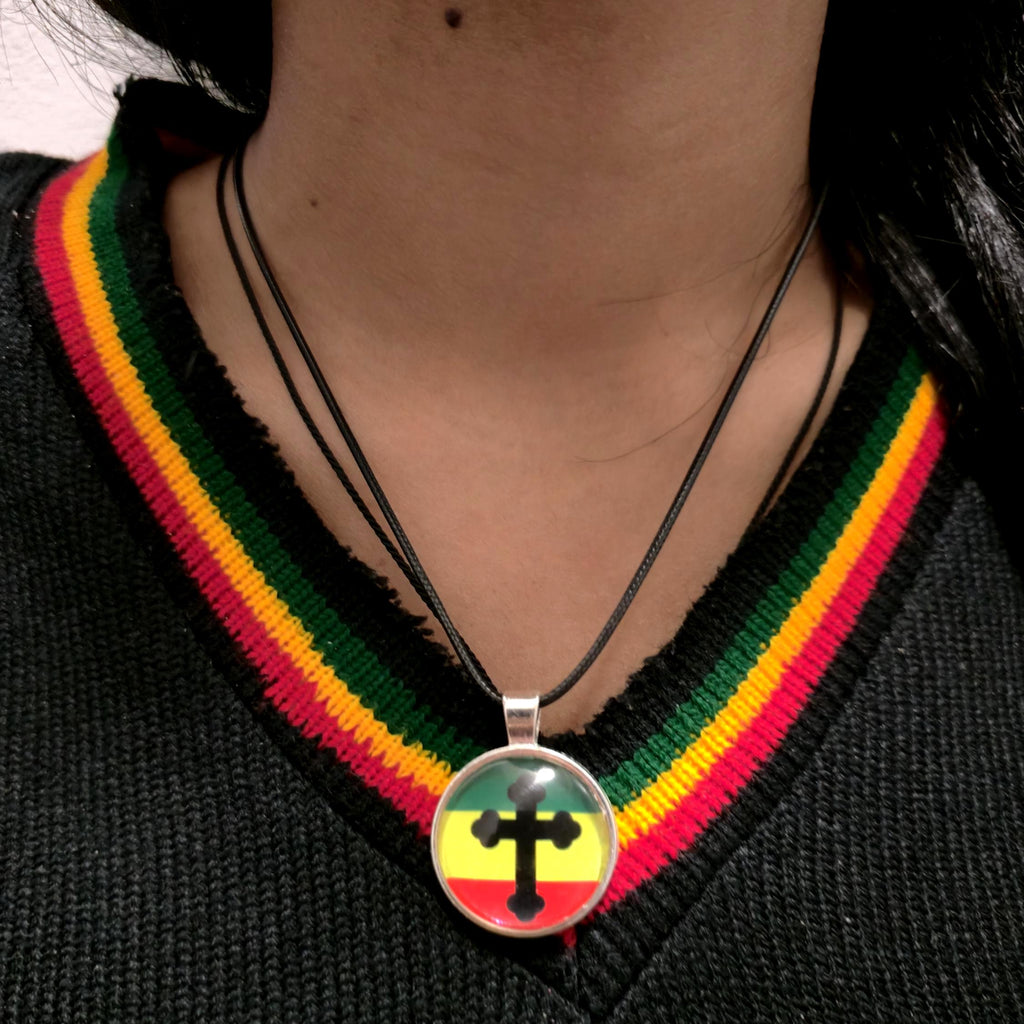 A close-up of a person wearing a black V-neck sweater with a green, yellow, and red striped collar trim, wearing a black cord necklace with a round pendant featuring a black cross over the Rastafarian flag colors.