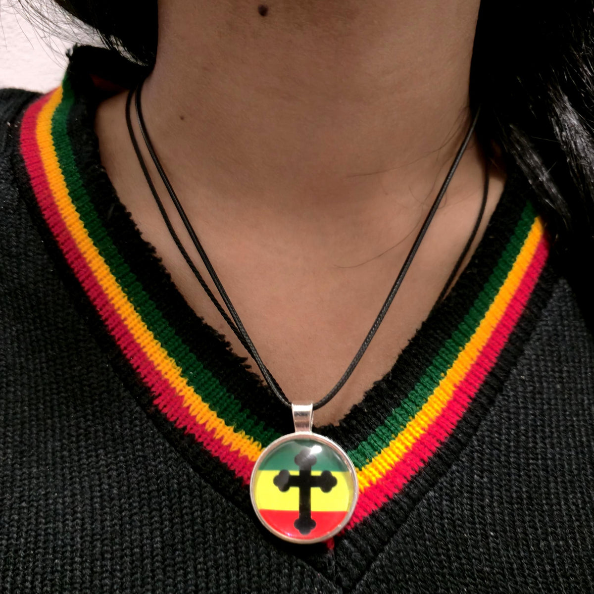 A close-up of a person wearing a black V-neck sweater with a green, yellow, and red striped collar trim, wearing a black cord necklace with a round pendant featuring a black cross over the Rastafarian flag colors.
