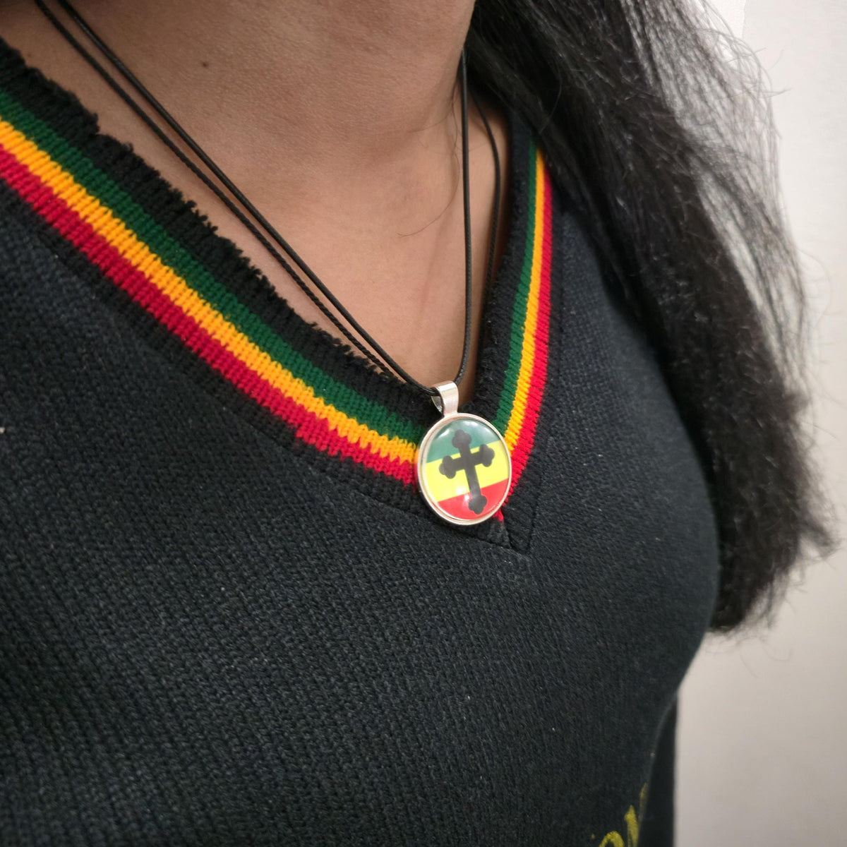 A close-up of a person wearing a Rastafarian-themed necklace with a green, yellow, and red striped collar on a black sweater. The necklace features a round pendant with a cross design in a Rastafarian flag color scheme.