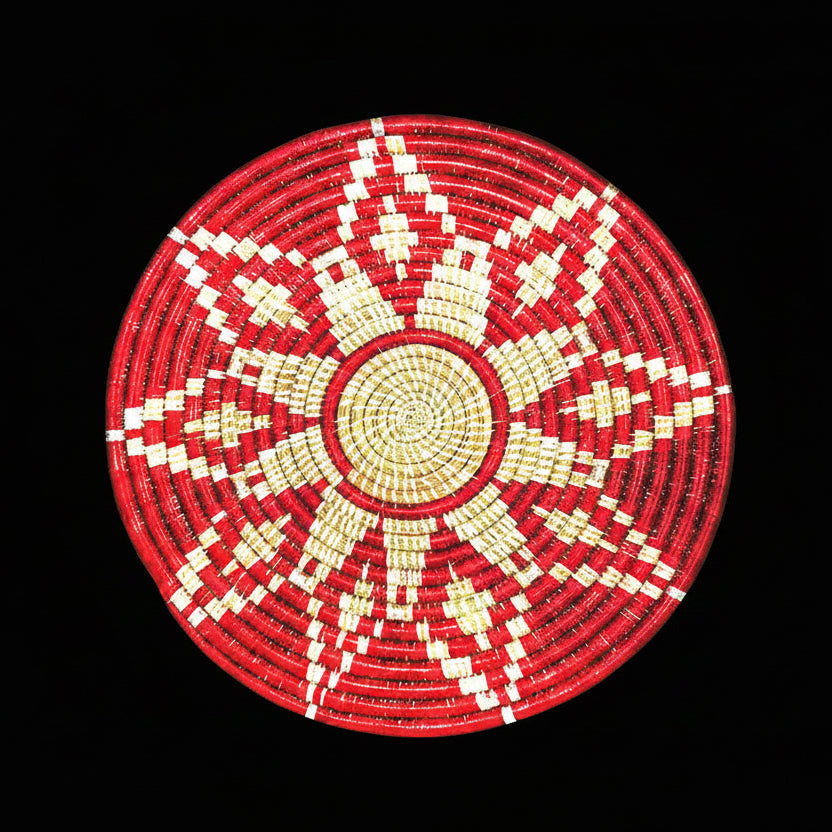 A circular hand-woven basket with a geometric starburst pattern in red and cream-white, isolated on a white background.