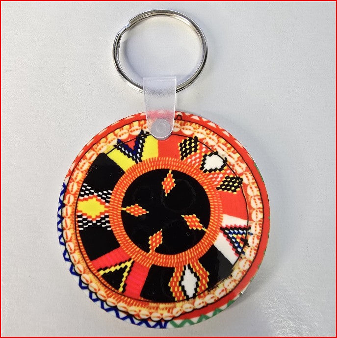 A round, handcrafted keychain featuring a vibrant, multicolored geometric pattern with orange, red, black, yellow, and white details, attached to a silver metal key ring with a clear plastic connector.