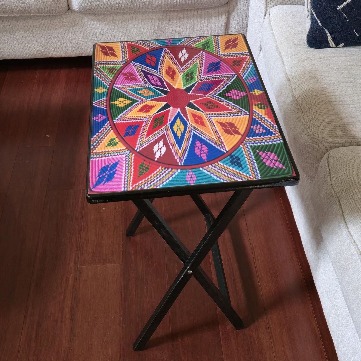 A square folding side table with a black metal frame and a vibrant, multicolored geometric patterned top, shown in a living room setting next to a white sofa on a wooden floor.