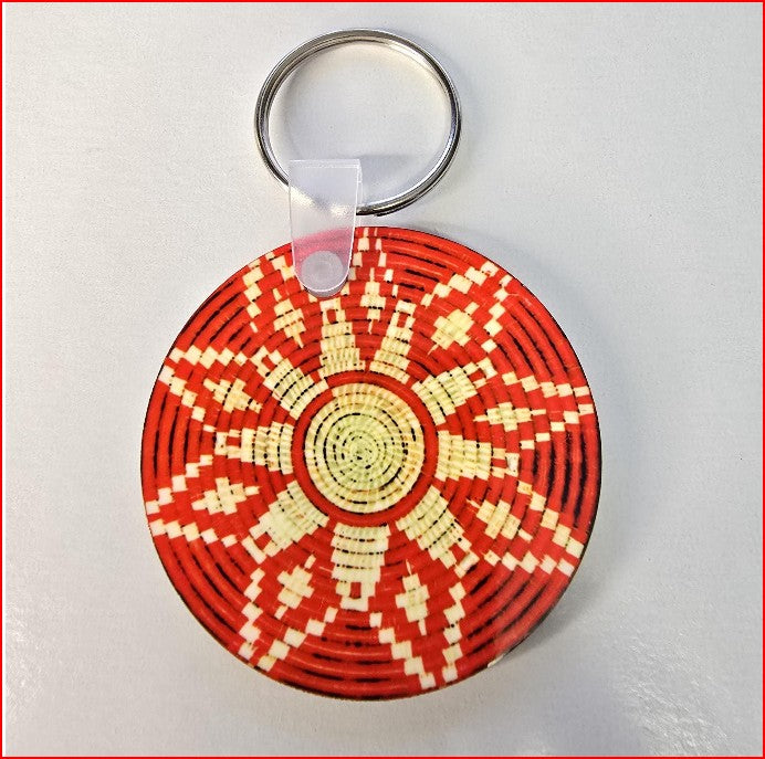 A round woven basket-style keychain with a red and cream geometric pattern, attached to a silver metal key ring with a clear plastic clip, shown on a light gray background.