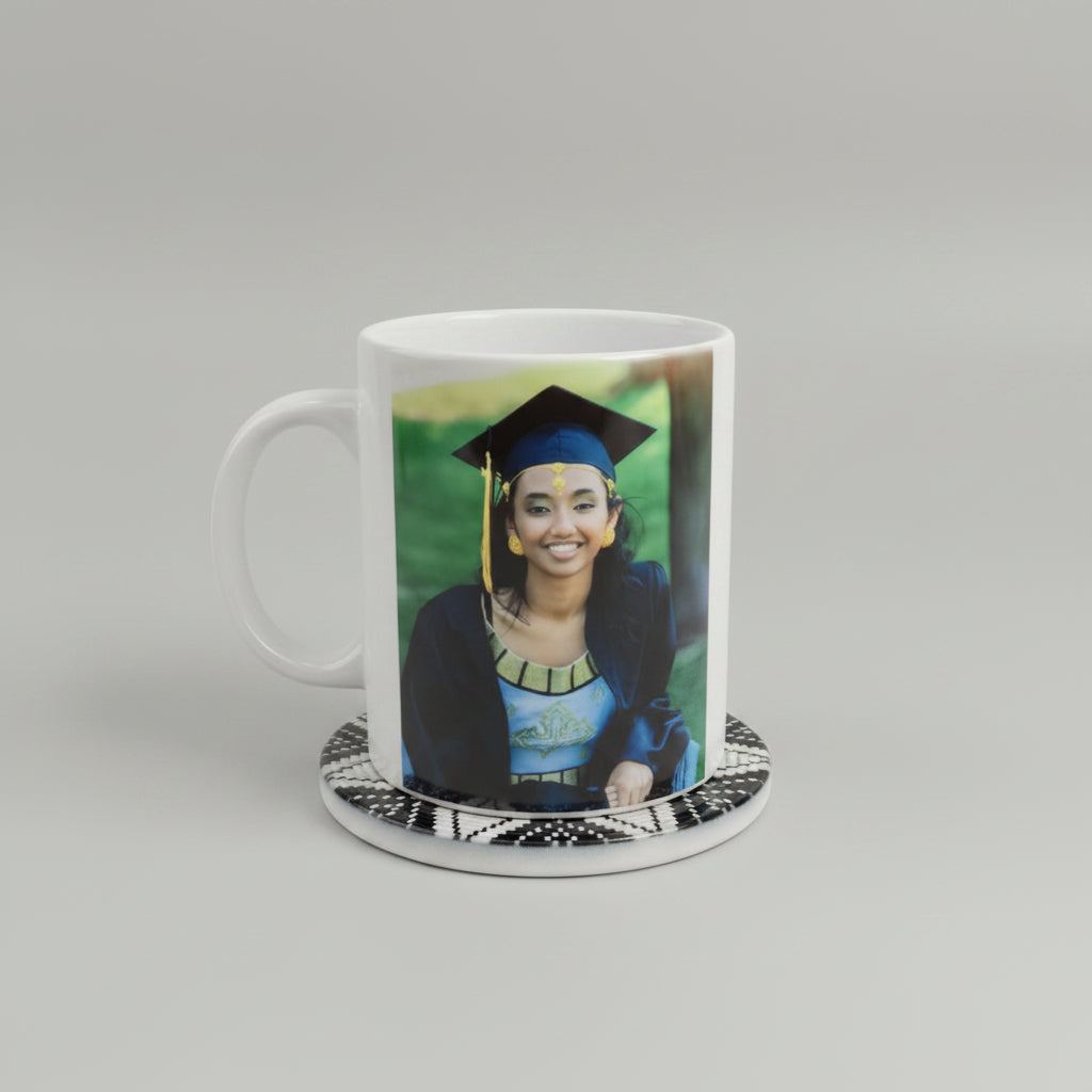 A white ceramic mug featuring a printed photo of a smiling woman in a graduation cap and gown, resting on a black and white patterned coaster against a light beige textured background.