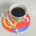 A white ceramic cup filled with black coffee sits on a matching saucer and spoon. The cup and spoon have a colorful illustrated border, while the saucer is red, yellow, and blue with a yellow emblem.