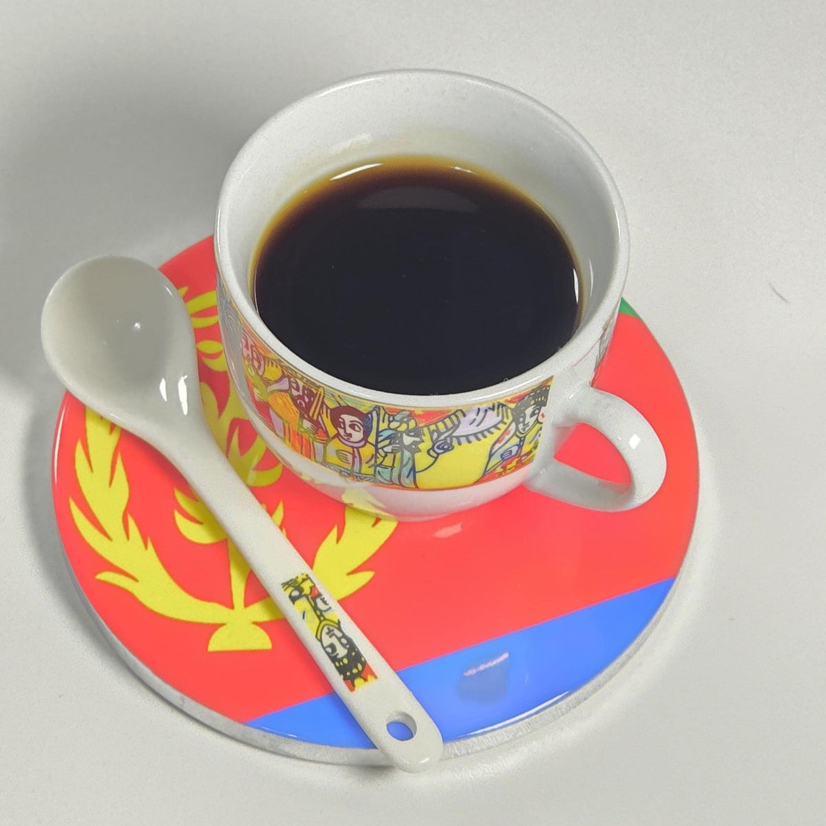 A white ceramic cup filled with black coffee sits on a matching saucer and spoon. The cup and spoon have a colorful illustrated border, while the saucer is red, yellow, and blue with a yellow emblem.