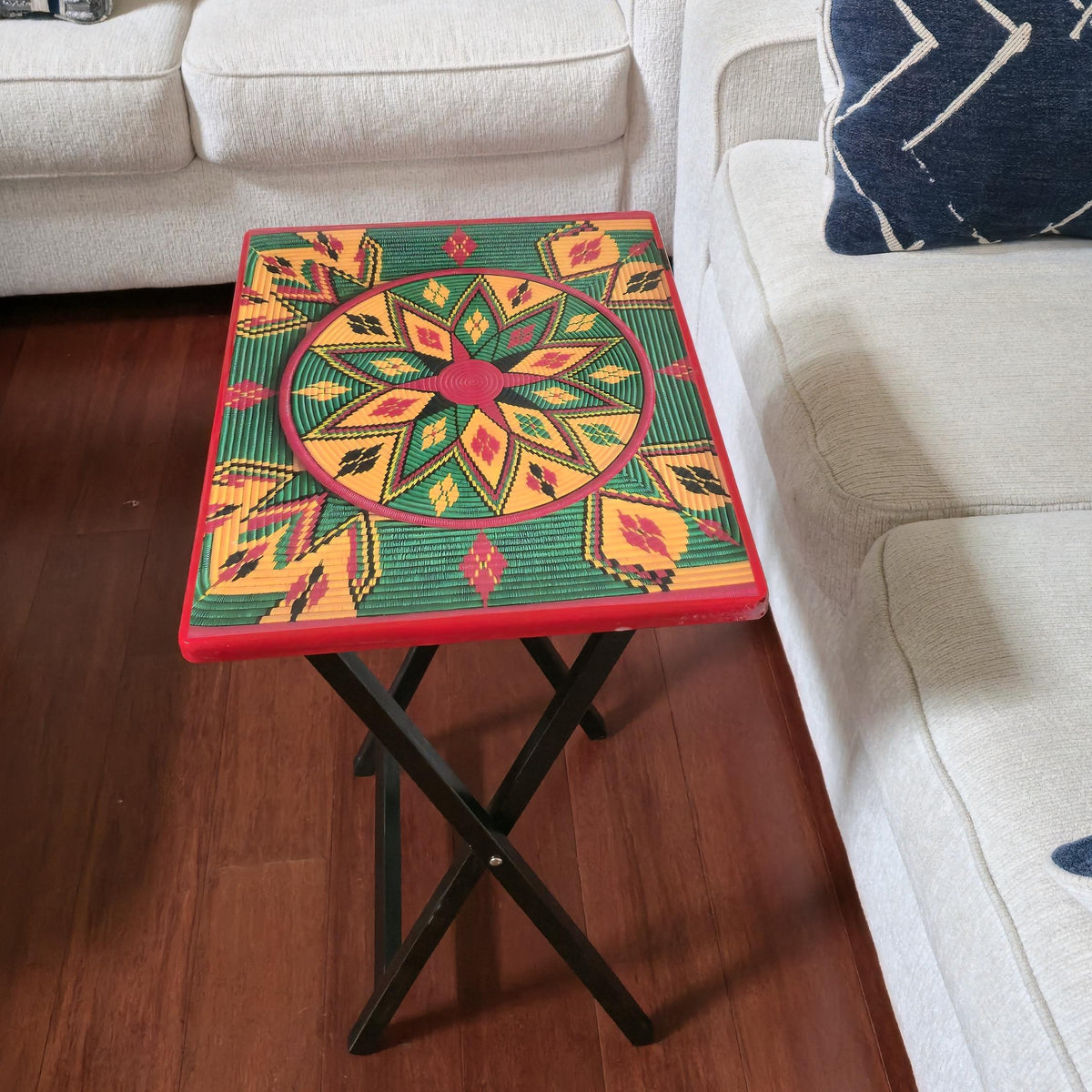 A square folding side table with a colorful geometric patterned top and black metal legs, placed in a living room between two white sofas on a wooden floor.