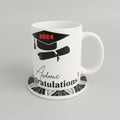 A white ceramic graduation mug printed with a black mortarboard cap, rolled diploma, and the words '2024' and 'Congratulations!' in black and red. The mug sits on a black-and-white patterned coaster on a beige textured surface against a neutral beige wall.