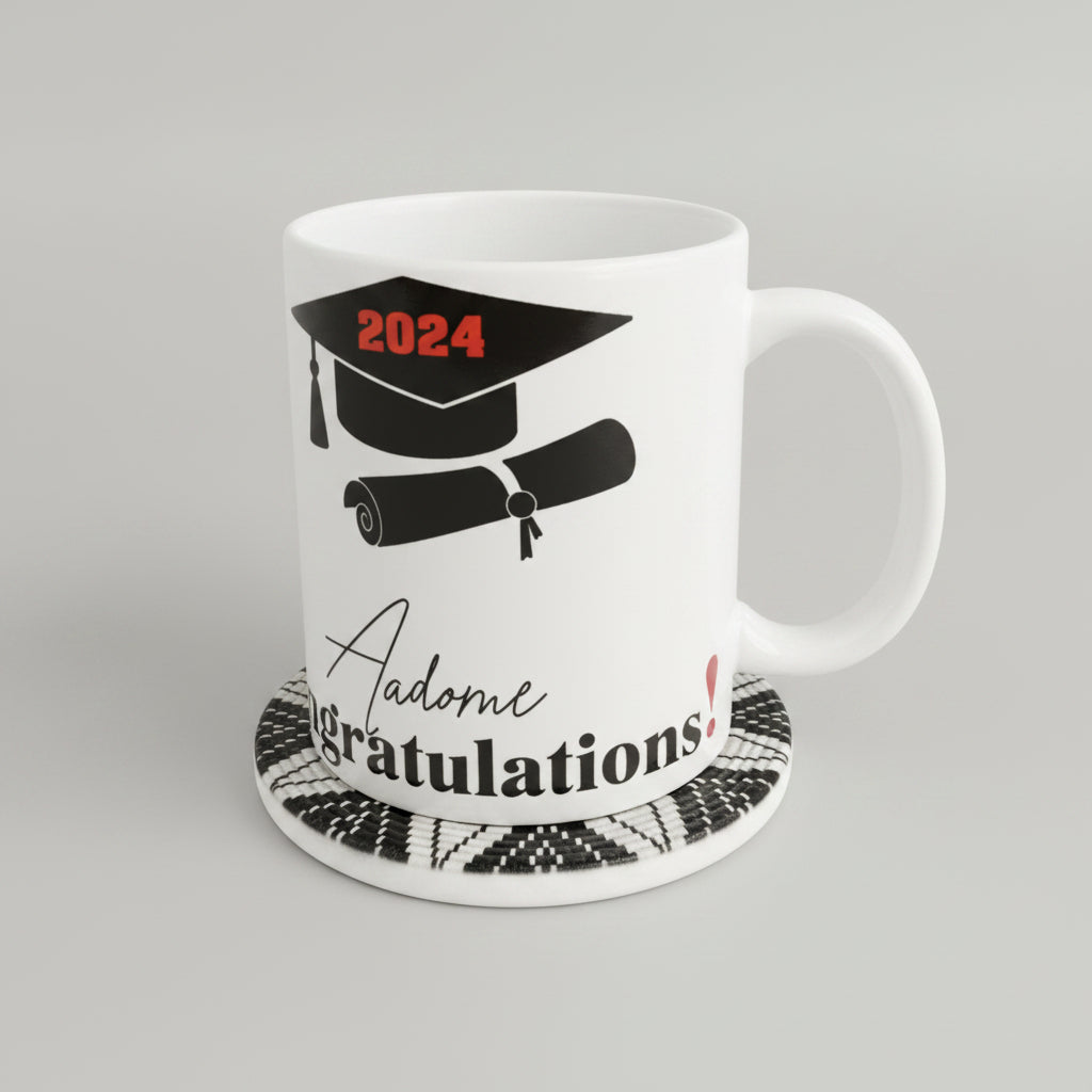 A white ceramic graduation mug printed with a black mortarboard cap, rolled diploma, and the words '2024' and 'Congratulations!' in black and red. The mug sits on a black-and-white patterned coaster on a beige textured surface against a neutral beige wall.
